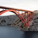 A striking red arch bridge spans a rocky landscape over a calm waterway, showcasing modern engineering.